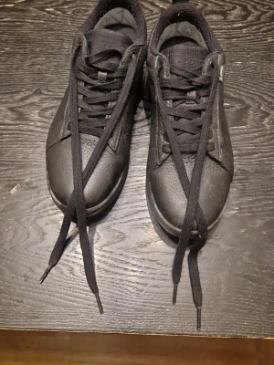 Svarta sneakers från Lacoste - Stilrena svarta sneakers från Lacoste i läder med klassisk låg profil och svarta skosnören. Vattentäta läder och gore tex material. Diskret logga på plösen och sula med rutmönster. Perfekta för en clean och enkel look.(Pris går att ändra)