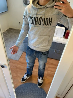 Grå HOODRICH hoodie med tryck - Snygg grå hoodie från HOODRICH med stor svart logga över bröstet och armarna. Hoodien har huva med dragsko och en klassisk magficka. Tillverkad i mjukt material som känns skönt mot huden. Perfekt för en avslappnad streetwear-look. Har två slitningar syns på det sista två bilderna