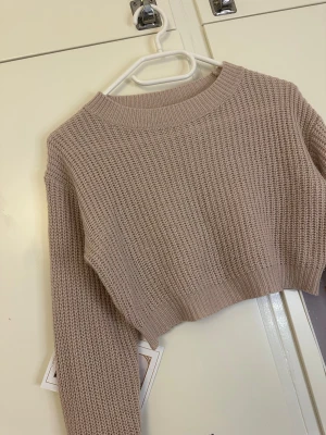 Beige cropped stickad tröja - Säljer en beige cropped stickad tröja med grov struktur och rund hals. Tröjan har långa ärmar och bred ribbad mudd vid hals, ärmslut och nederkant. Perfekt för lager på lager och enkel att matcha med olika stilar.