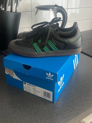 Adidas Samba OG svart/grön sneakers - Klassiska Adidas Samba OG sneakers i svart och grå mocka med gröna ränder på sidorna och brun gummisula. Skorna har lågt skaft, svarta skosnören och ikonisk retrostil. Perfekta för dig som gillar streetwear och tidlös design.