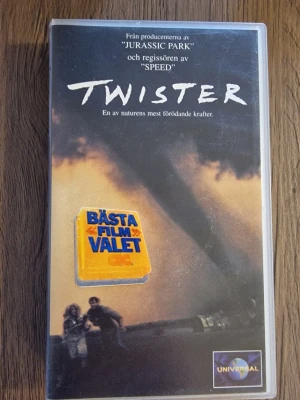Twister VHS-film från Universal - Twister på VHS, actionfylld katastroffilm från 1996. Omslaget är svart och gult med en dramatisk tornado och texten 'Bästa filmvalet'. Filmen är på svenska med svensk text och har klassiska 90-talsvibbar. Plastfodral med tryckt omslag och Universal-logga.