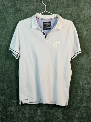 Ljusblå pikétröja från Bondelid - Klassisk ljusblå pikétröja från Bondelid med vit broderad logga och texten B.D.L. på bröstet. Tröjan har krage, knappar i halsen och vita detaljer på ärmsluten. Perfekt till en avslappnad men snygg stil.