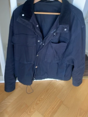 Svart overshirt jacka med fickor från Zara - Svart overshirt jacka från Zara med flera stora fickor framtill, dragkedja och tryckknappar. Jackan har en krage i manchester och justerbar dragsko nedtill. Perfekt för dig som gillar en avslappnad och funktionell stil.