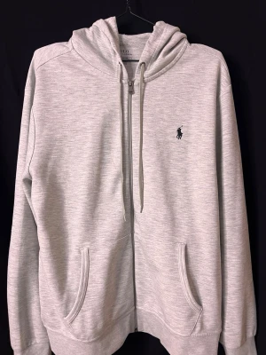 Ljusgrå hoodie från Polo Ralph Lauren - Ljusgrå hoodie från Polo Ralph Lauren med dragkedja, två fickor framtill och klassisk huva med dragsko. Ikoniska Polo-loggan broderad på bröstet. Tillverkad i mjukt bomullsmaterial som känns skönt mot huden. Perfekt för en avslappnad och stilren look. Skick 9/10