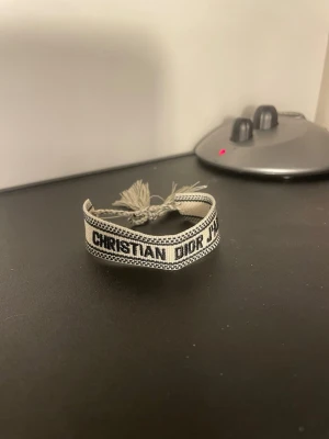 Christian Dior J'Adior tygarmband - Snyggt beige och svart tygarmband från Christian Dior med texten 'J'Adior'. Armbandet har flätade band och tofsar i ändarna för en cool och avslappnad look. Perfekt accessoar för att lyfta din outfit med en lyxig touch.