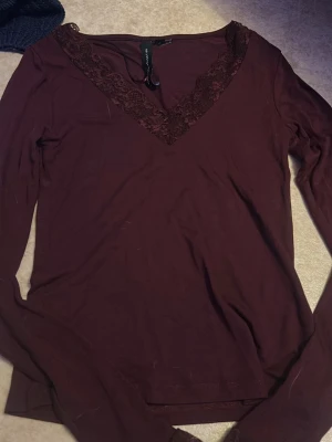 Mörkröd topp med spets  - Snygg mörkröd långärmad topp med v-ringning och dekorativ spets runt halsen. Perfekt att styla med jeans eller kjol för en trendig look. Materialet känns mjukt och stretchigt.