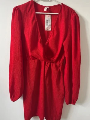 Röd omlottklänning med v-ringning - Trendig röd kortklänning med omlott och djup v-ringning. Klänningen har långärmade puffärmar och är gjord i ett strukturerat tyg som ger en cool look. Perfekt för dig som vill sticka ut med färg och stil.