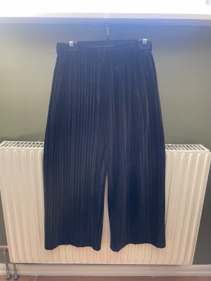 Svarta utsvängda byxor  - Säljer ett par svarta utsvängda byxor i mjuk sammet med  resår i midjan. De har raka ben och en lyxig känsla som passar perfekt till din outfit. Materialet är glansigt och ger en snygg look.