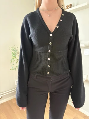 Svart ribbad kofta med v-ringning - Stilren svart kofta med v-ringning och ribbad midja. Koftan har långa ärmar och knäpps med vita knappar framtill. Passformen är figurnära vid midjan och lösare vid ärmarna, vilket ger en trendig siluett.