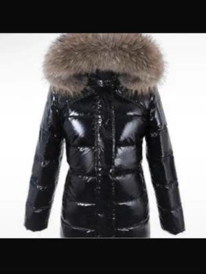 Svart glansig dunjacka med fuskpäls - Snygg svart dunjacka med glansigt material och stor huva kantad med fluffig fuskpäls. Jackan har quiltad design och går ner över höfterna för extra värme. Perfekt för kalla dagar när du vill hålla stilen.