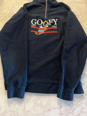 Mörkblå Goofy fleece från Disney - Mysig mörkblå fleece med Goofy-brodyr och texten 'GOOFY EST. 1932' på bröstet. Tröjan har halv dragkedja framtill och långa ärmar. Perfekt för dig som gillar Disney. 