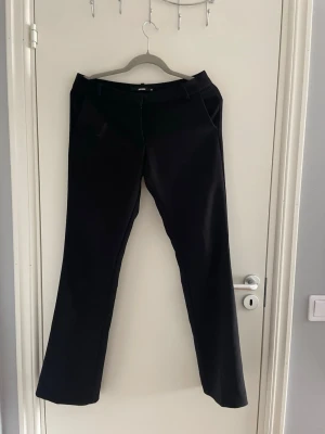 Svarta kostymbyxor med utsvängdaben  - Stilrena svarta kostymbyxor med utsvängda ben och diskreta fickor fram. Byxorna har hällor för skärp och klassisk dragkedja. Perfekta för en clean och dressad look. Materialet ser ut att vara en mjuk och lite stretchig blandning. Som man ser på sista bilden har en flärp gått av men de är lätt fixat💘