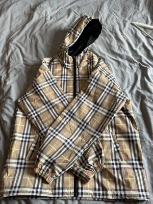  beige rutig vindjacka - Snygg beige vindjacka  med klassiskt rutmönster i svart, vitt och gult. Jackan har huva, dragkedja framtill och två sidofickor. Materialet är lätt och vattenavvisande, perfekt för blåsiga dagar. Elastiska muddar vid ärmsluten för extra komfort. det står XXXL men den passar XL