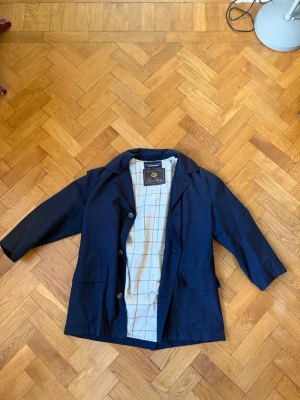 Loro Piana jacka  - Klassisk mörkblå rock från Loro Piana med rutigt innerfoder i vitt, rött och blått. Rocken har tre knappar framtill, två stora fickor och en stilren krage. Perfekt för dig som vill ha en tidlös och snygg look. Gjord i en ull blandning. Pytte pytte lite smutsig på en ficka, går lätt bort i tvätten. Kort i armarna. 