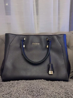 Svart handväska från Guess - Stilren svart handväska från Guess i konstläder med guldfärgade detaljer och logotyp framtill. Väskan har två handtag, dekorativ hängare med G-detalj och ljusrosa innerfoder med mönster. Perfekt för dig som vill ha en klassisk och rymlig väska.