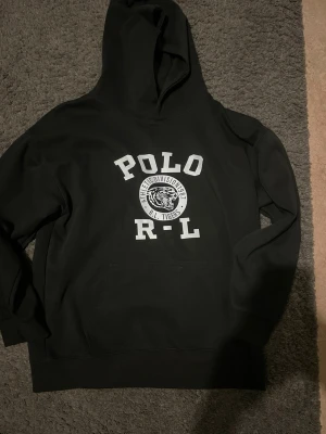 Polo Ralph Lauren hoodie  - Svart hoodie från Polo Ralph Lauren med stor vit logga och texten 'POLO R-L' samt en tiger på bröstet. Klassisk känguruficka framtill och huva. Tillverkad i mjukt bomullsmaterial, perfekt för en avslappnad streetwear-look.