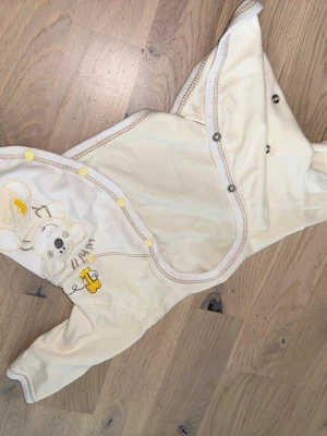 Krämvit omlottbody med nallemotiv - Krämvit omlottbody med nallemotiv Supersöt krämvit omlottbody för baby med broderat nallemotiv och gula detaljer. Tryckknappar längs sidan och ärmar för enkel påklädning. Mjuk bomullskänsla och fina kontrastsömmar. Perfekt för mysiga dagar hemma. Omlottbody med långa ärmar och tryckknappar framtill. Bodyn har ett broderat motiv av en björn med en honungsburk och en bi på ena sidan.cirka 0-5 mån 