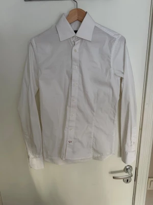 Klassisk vit skjorta med knappar - Säljer en stilren vit skjorta med lång ärm och klassisk krage. Skjortan har knäppning framtill och är tillverkad i bomull, perfekt för en clean och enkel look. Passar till många olika outfits och är ett måste i garderoben.