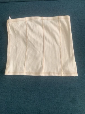 Beige ribbad tubtopp - Säljer en beige ribbad tubtopp med sömdetaljer framtill. Toppen är stretchig och har en enkel, stilren design utan axelband. Perfekt att matcha med jeans eller kjol för en trendig look. Storlek L 