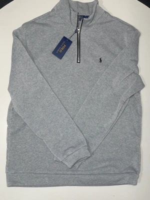 Grå halvzip hoodie från Polo Ralph Lauren - Snygg grå halvzip hoodie från Polo Ralph Lauren med klassisk broderad logga på bröstet. Tillverkad i mjukt sweatshirtmaterial och har hög krage med dragkedja. Perfekt för en avslappnad och stilren look. Size är M men liknar Xl och L den är inte så lite som en M