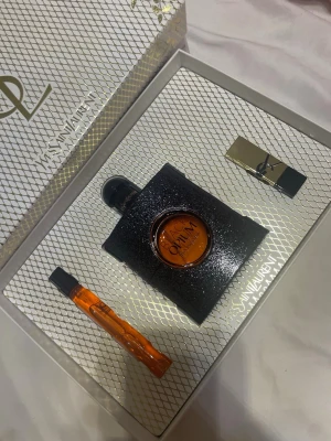 Yves Saint Laurent Black Opium presentset - Lyxigt presentset från Yves Saint Laurent med Black Opium parfym i svart glittrig flaska, matchande miniflaska och ett elegant läppstift i guld och svart. Allt levereras i en exklusiv ask med guldmönster och YSL-logga.