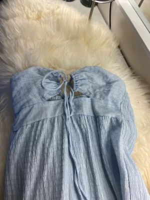 Ljusblå maxiklänning med knyt - Supersöt ljusblå maxiklänning med smockad struktur och knyt fram vid bysten. Klänningen har tunna axelband och faller luftigt ner till anklarna. Perfekt för varma dagar och ger en chill sommarkänsla.