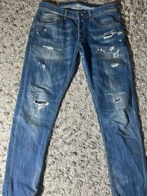 Blå dondup jeans med schyssta slitningar skinny fit - Säljer ett par blå jeans med smal passform och slitna detaljer samt hål på benen. Jeansen har klassisk femficksmodell och knappgylf. Perfekta för en avslappnad och trendig look. Se defekt på andra bilden. Går dock lätt att laga och eftersom att jeansen har slitningar ser det bara ut som en fet detalj 