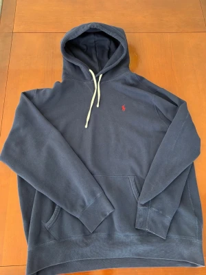 Mörkblå hoodie från Polo Ralph Lauren - Snygg mörkblå hoodie från Polo Ralph Lauren med klassisk huva och vita dragsnören. Har en liten röd logga broderad på bröstet och en stor magficka framtill. Tillverkad i mjuk bomullsblandning som är perfekt för chill dagar.