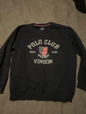 Svart tröja från Vinson Polo Club - Snygg svart tröja från Vinson Polo Club med broderad text och amerikansk flagga på bröstet. Rund halsringning och långa ärmar. Perfekt för en avslappnad och sportig stil.