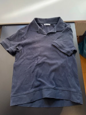 Mörkblå ribbad t-shirt från Zara - Snygg mörkblå t-shirt från Zara med ribbad struktur och klassisk krage. T-shirten har korta ärmar och är tillverkad i ett mjukt material som känns skönt mot huden. Perfekt för en stilren och avslappnad look.