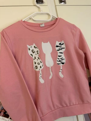 Rosa sweatshirt med kattmotiv - Söt rosa sweatshirt med tryck av tre katter på bröstet, där två har djurmönster i svart och beige. Tröjan har rund halsringning och långa ärmar. Perfekt för dig som gillar gulliga och lekfulla plagg.