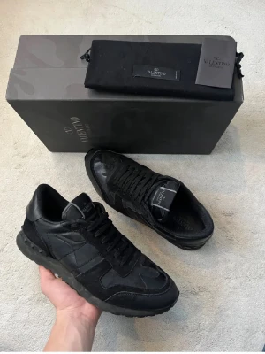 Svarta Valentino Garavani sneakers - Snygga svarta sneakers från Valentino Garavani med diskret camomönster och detaljer i mocka och läder. Klassisk låg modell med svarta skosnören och robust sula. Perfekta för dig som vill ha en exklusiv och stilren look. 