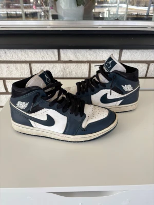 Air Jordan 1 Mid Navy/Vit - Snygga Air Jordan 1 Mid sneakers i marinblått och vitt läder med svarta skosnören och klassisk Jumpman-logga på plösen. Ikonisk vinge på sidan och perforerad tå för extra stil. Perfekta för dig som gillar streetwear och vill sticka ut.