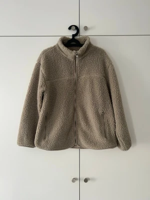 Beige teddyjacka med dragkedja - Mysig beige teddyjacka i storlek M med hög krage och hel dragkedja framtill. Jackan har två sidofickor med dragkedja och är gjord i fluffigt teddy-material som håller dig varm. Perfekt för kyliga dagar och enkel att matcha med olika outfits.