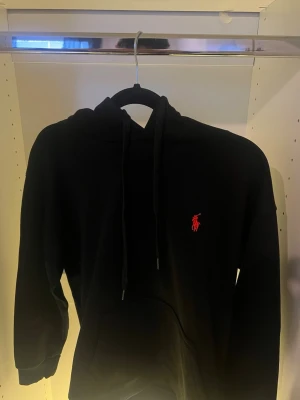 Svart hoodie från Polo Ralph Lauren -  Jag säljer den eftersom den var för liten för mig!Svart hoodie från Polo Ralph Lauren med klassisk röd logga broderad på bröstet. Hoodien har huva med dragsko och en stor magficka. Tillverkad i mjukt bomullsmaterial som känns skönt mot huden. Perfekt för en avslappnad och stilren look.