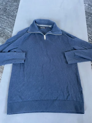 Blå halvzip tröja från Jack & Jones - Mörkblå stickad tröja från Jack & Jones med halv dragkedja och hög krage. Tröjan har ribbade muddar vid ärmslut och nederkant, perfekt för en avslappnad men snygg stil. Materialet känns mjukt och bekvämt. Hör av er vid funderingar! 😁