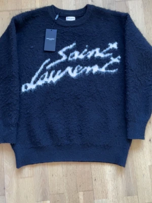 Svart Saint Laurent stickad tröja - Snygg svart stickad tröja från Saint Laurent med vit logotyp i stor text framtill. Tröjan har en fluffig struktur, rund halsringning och ribbade muddar vid ärmslut och nederkant. Perfekt för dig som vill ha en trendig och exklusiv look.