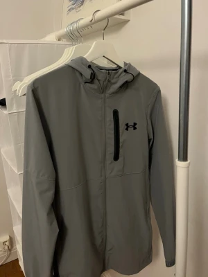 Grå vindjacka från Under Armour - Snygg grå vindjacka från Under Armour. Den är i toppskick och använts typ 5 gånger. Den är i storlek S men passar även storlek M. 