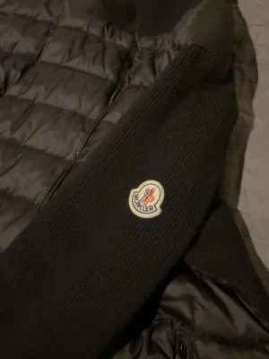 Svart cardigan från Moncler - Snygg svart cardigan från Moncler med quiltad kropp och stickade ärmar. Jackan har dragkedja framtill, ståkrage och Moncler-logga på ena ärmen. Perfekt för kalla vinterdagar och har en modern, stilren look. 