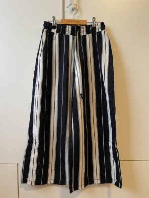Randiga vida byxor svart/vit - Säljer ett par snygga byxor med svarta och vita vertikala ränder. Byxorna har resår i midjan med dragsko och vida ben för en avslappnad look. Perfekta för sommaren och riktigt sköna att bära. Modellen är tänkt att vara kortare och sluta ovanför ankeln.