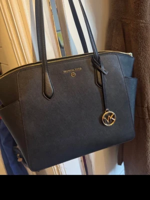 Svart Michael Kors handväska - Snygg svart handväska från Michael Kors i tåligt saffiano-läder. Väskan har guldfärgade detaljer, dragkedja upptill och ett hängande MK-emblem. Invändigt finns fack och fickor för bra organisering. Perfekt accessoar för dig som gillar stilrena och klassiska väskor. Givetvis äkta o äkthetsbevis finns i väskans fack. ♥️lappen sitter kvar inuti, endast testad sen hängt i garderoben 1år. 