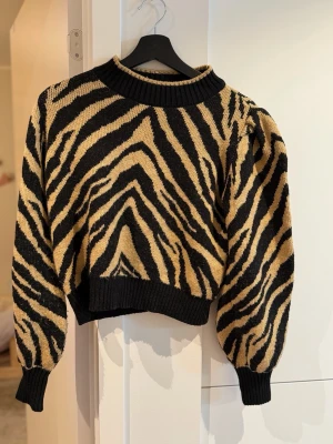 Zebramönstrad stickad tröja - Cool stickad tröja med zebramönster i svart och beige. Tröjan har ballongärmar, ribbade muddar och en rund halsringning. Perfekt för dig som vill sticka ut med ett djurinspirerat plagg.