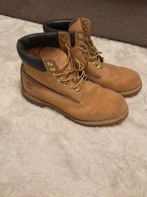 Timberland klassiska kängor beige - Säljer ett par klassiska Timberland kängor i beige nubuckläder med robust gummisula och svart vadderad kant runt ankeln. Kängorna har metallöljetter och kraftiga snören, perfekt för dig som vill ha en ikonisk streetwear-look.
