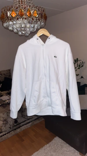 Vit hoodie från Lacoste med dragkedja - Vit hoodie från Lacoste med klassisk krokodillogga på bröstet. Tröjan har huva med snörhål, två fickor framtill och hel dragkedja. Tillverkad i mjuk bomull som känns skön mot huden. Perfekt för chill dagar och enkel att matcha med jeans eller joggers.