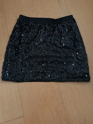 Low waist paljettkjol - Passar en xs! Snygg svart mini kortkjol från Lindex täckt med glittrande paljetter. Kjolen har en elastisk midja och är perfekt för dig som vill sticka ut med en festlig look. Passar dig som gillar att glänsa och vill ha något extra till outfiten. Den är low waist och man kan även vika in kanterna om man vill ha ännu mer low waist.
