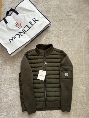 Olivgrön pufferjacka från Moncler - Snygg olivgrön pufferjacka från Moncler med quiltad front och ärmar i stickad struktur. Jackan har hög krage, dragkedja framtill och en ficka med Moncler-logga på ena ärmen. Perfekt för kalla dagar och riktigt trendig look.