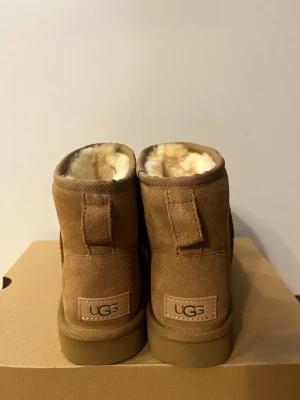 Bruna UGG Classic Mini boots - Säljer ett par bruna UGG Classic Mini boots i mocka med mjukt, vitt fårskinnsfoder. Skorna har en klassisk rund tå och grov sula i gummi. Stl 36, helt oanvända!