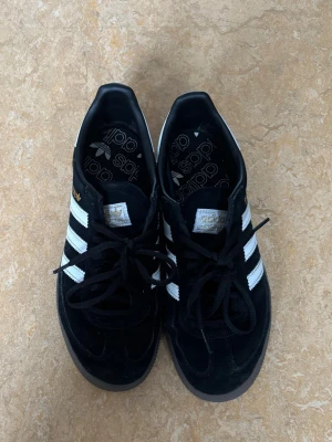 Adidas Spezial svarta sneakers - Svarta Adidas Spezial sneakers med klassiska vita ränder på sidorna och gummisula. Skon har gulddetaljer vid loggan och är tillverkad i mocka och textil. Perfekt för alla tillfällen 