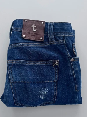 Blå jeans från Tramarossa - Säljer ett par snygga blå jeans från Tramarossa med klassisk femficksmodell och knappgylf. Jeansen har en cool sliten effekt på ena benet och bakfickan, samt detaljer i metall och läderpatch med logga bak. Perfekta för dig som gillar stilrena men ändå unika jeans.