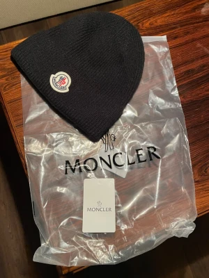 Svart Moncler mössa i ull - Svart ribbstickad mössa från Moncler med klassisk logotyp framtill. Tillverkad i mjuk ull och levereras med originalpåse och tagg. Perfekt accessoar för kyliga dagar och en stilren detalj till din outfit.